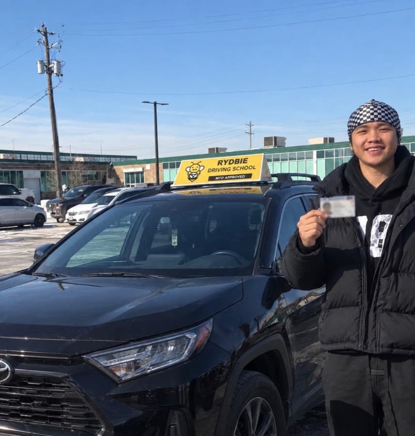 A Rydbie student holding their new Ontario driver's licence beside a Rydbie car